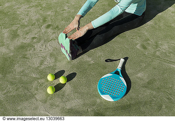 Low section of woman stretching with tennis racket and balls on court during sunny day