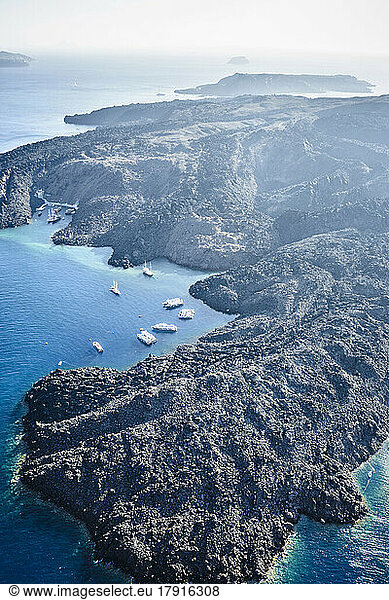 Luftaufnahme der Landschaft einer Insel in der Caldera  vulkanisches Gestein und steile Klippen sowie geschützte Anlegestellen mit Booten.