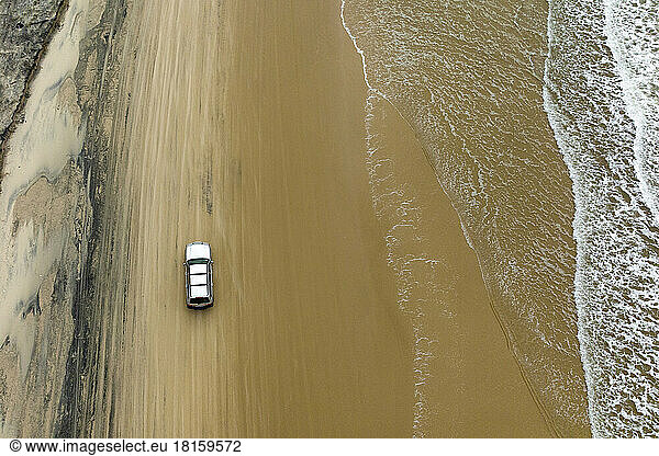 Luftaufnahme eines Geländewagens  der auf einer Strandstraße mit Wellen geparkt ist