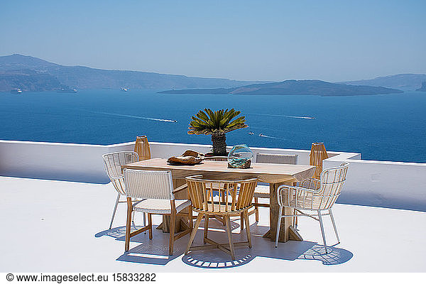 Möbel  bestehend aus einem Tisch und einigen Stühlen  auf einer weißen Terrasse eines Hauses in Santorin in Griechenland  wo man das Essen genießen kann  während man eine romantische Meereslandschaft bis hin zum blauen Ägäischen Meer sieht. Horizontal ph