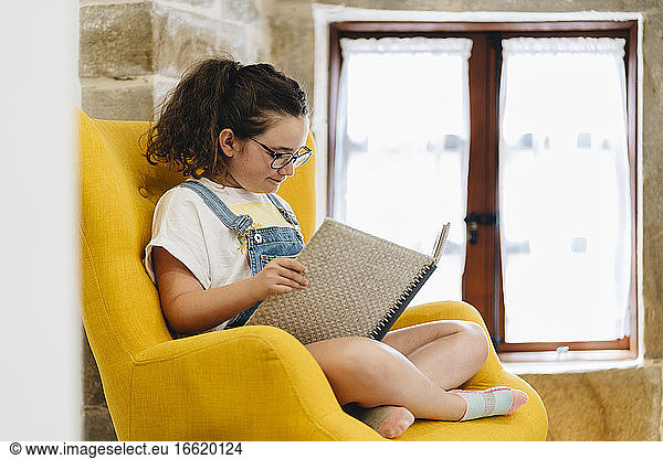 Mädchen liest ein Buch  während sie zu Hause auf einem Sessel sitzt