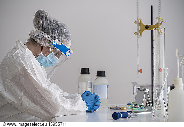 Müde Wissenschaftlerin in Schutzkleidung am Schreibtisch sitzend