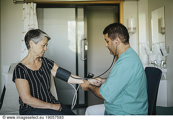 Männlicher Arzt  der im Krankenhaus sitzend den Blutdruck einer älteren Frau kontrolliert