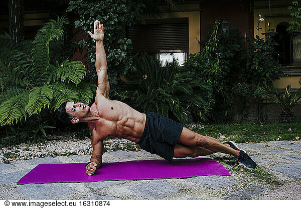 Männlicher Athlet ohne Hemd übt die Plank-Position auf einer Matte gegen Pflanzen im Hof
