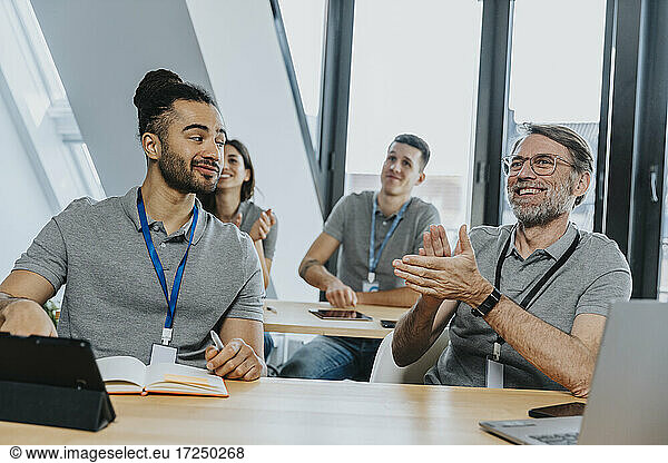 Männlicher Auszubildender schaut auf einen lächelnden  klatschenden Kollegen  während er auf einer Bank in einer Schulklasse sitzt