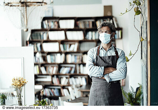 Männlicher Café-Besitzer mit Gesichtsschutzmaske steht mit verschränkten Armen in einem Café