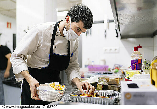 Männlicher Koch mit Schutzmaske ordnet Pommes frites in einem Folienbehälter in einer Restaurantküche an