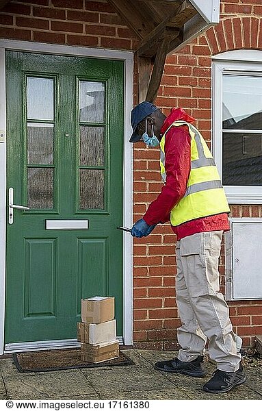 Männlicher Kurier  der während der Covid-19-Epidemie Pakete und Päckchen ausliefert  mit Handschuhen und Gesichtsmaske  Hampshire  England  Vereinigtes Königreich; Aufnahme der Auslieferung mit dem Mobiltelefon.