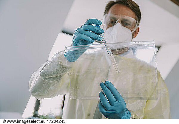 Männlicher Mitarbeiter im Gesundheitswesen  der im Untersuchungsraum stehend einen Nasenabstrich in einer Plastiktüte nimmt