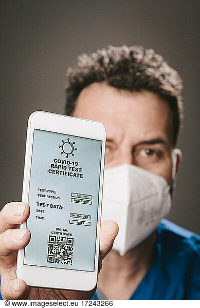 Männlicher Mitarbeiter im Gesundheitswesen mit Gesichtsmaske zeigt Schnelltest-Zertifikat auf einem Mobiltelefon