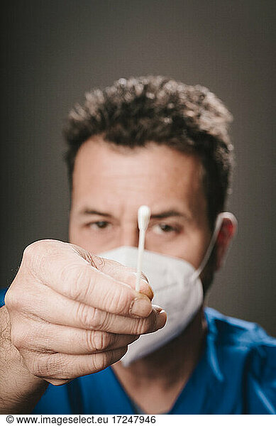 Männlicher Mitarbeiter im Gesundheitswesen mit Gesichtsschutzmaske  der einen Nasenabstrich vor einem grauen Hintergrund zeigt