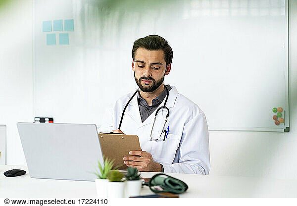 Männlicher Mitarbeiter im Gesundheitswesen mit Klemmbrett und Laptop am Schreibtisch sitzend