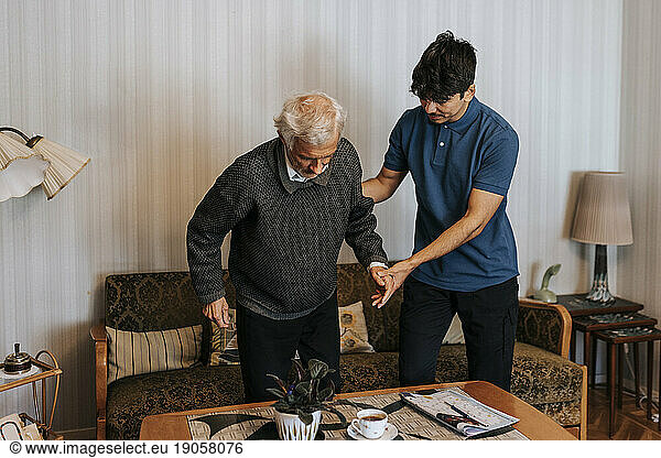 Männlicher Pflegehelfer unterstützt älteren Mann beim Aufstehen vom Sofa zu Hause