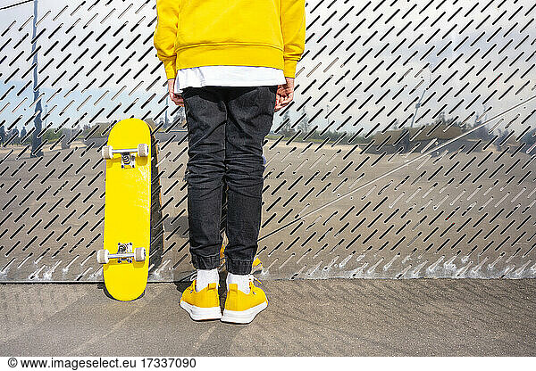 Männlicher Skater mit gelben Schuhen steht mit einem Skateboard vor einer Metallwand