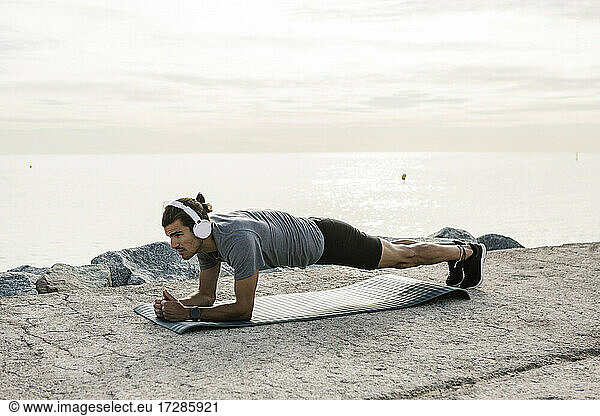 Männlicher Sportler mit Kopfhörern  der auf einer Trainingsmatte die Plank-Position übt