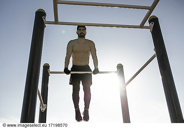 Männlicher Sportler ohne Hemd trainiert am Reck im Park gegen den klaren Himmel
