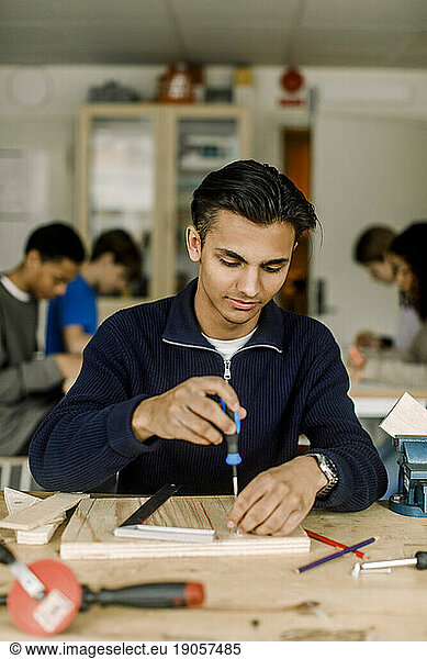 Männlicher Teenager  der einen Schraubenzieher auf Holz benutzt  während er in der High School im Tischlerkurs lernt