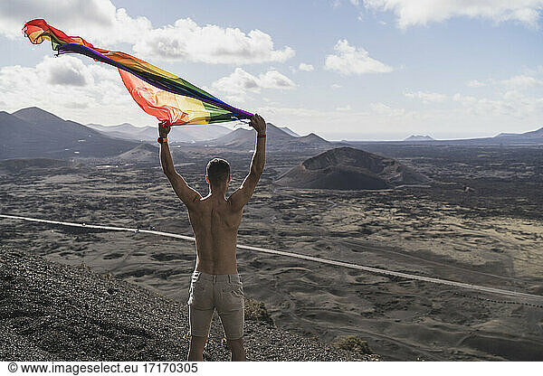 Männlicher Tourist ohne Hemd schwenkt LGBTQI-Flagge am Vulkan El Cuervo während der Ferien  Lanzarote  Spanien