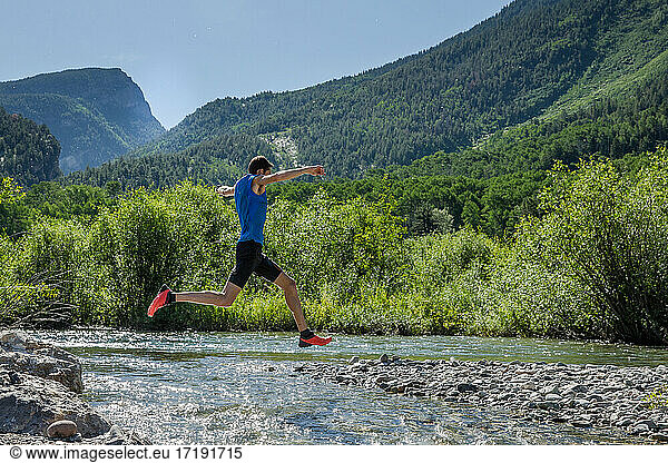 Männlicher Trailrunner springt an einem sonnigen Tag in den Bergen über einen felsigen Fluss