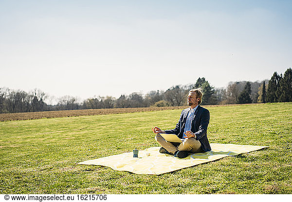 Männlicher Unternehmer sitzt mit Laptop und meditiert im Lotussitz auf einer Picknickdecke im Park gegen den klaren Himmel