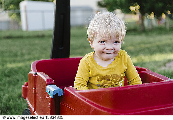 Männliches Kleinkind sitzt in einem roten Wagen im Garten der Familie.
