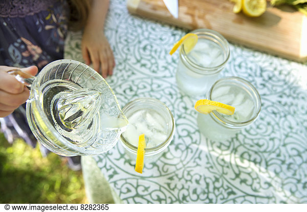 Making Lemonade. Overhead Shot Of Lemonade Glasses With A Fresh Slice Of Lemon In The Edge Of The Glass. A Child Pouring The Drink From A Jug.
