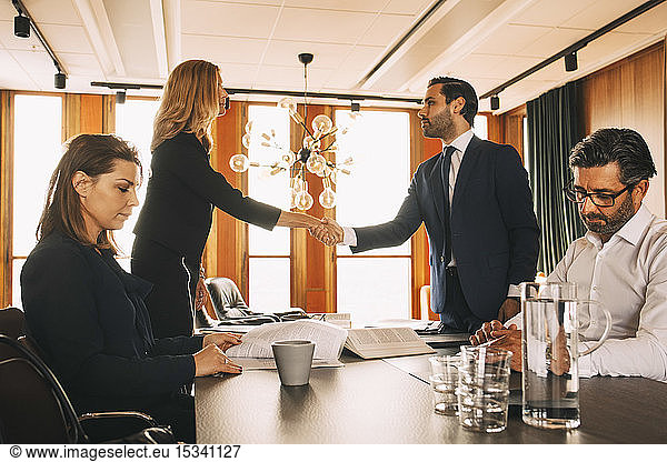 Male and female coworkers working while lawyers shaking hands at table in law office