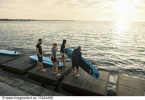 Male and female friends carrying paddleboard while standing on pier by sea