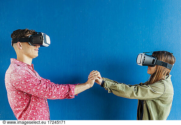 Male and female friends holding hands while standing by blue wall