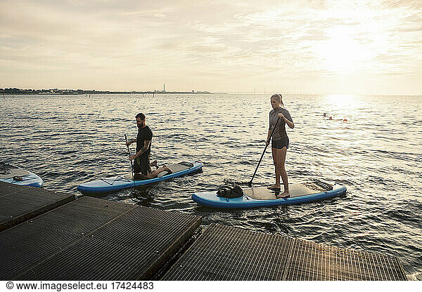 Male and female friends rowing paddleboard by pier in sea