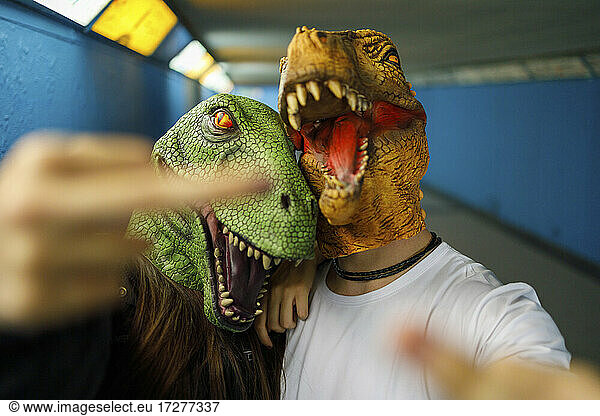 Male and female friends showing middle finger while wearing dinosaur mask in corridor
