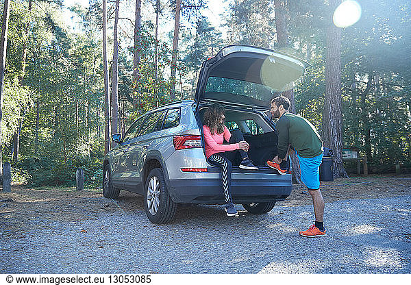 Male and female runners tying laces on car boot in sunlit forest