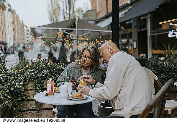 Male and female senior friends discussing over smart phone while having food at sidewalk cafe