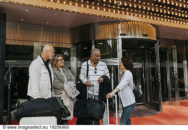 Male and female senior friends with luggage talking to each other while standing outside movie theater