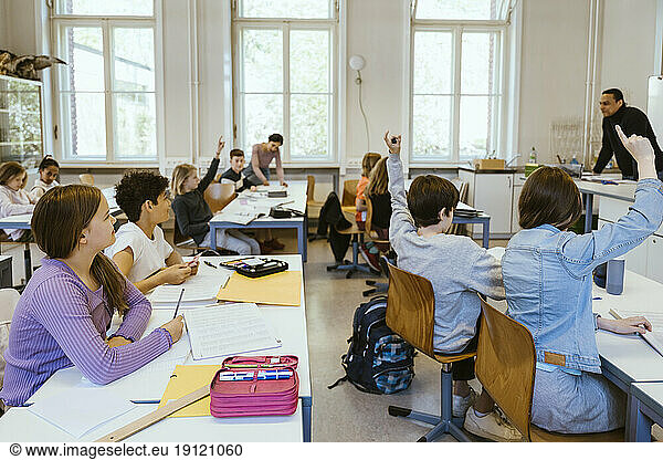 Male and female students raising hands while sitting on chairs in classroom