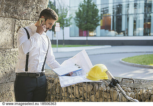 Male architect talking on mobile phone and looking blueprint at construction site  Munich  Bavaria  Germany