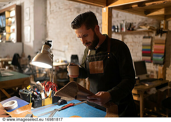Male artisan examining leather and drinking hot beverage