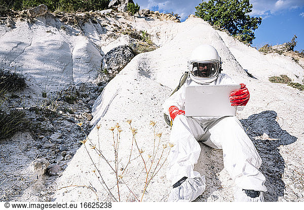 Male astronaut using laptop while sitting on mountain