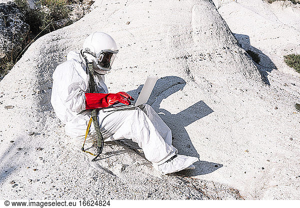 Male astronaut working over laptop while sitting on mountain
