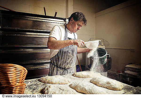 Male baker sprinkling flour on bread dough at bakery