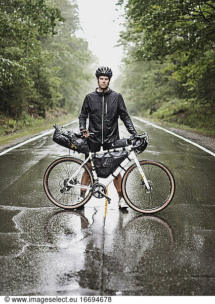 male bike packer stands with bike in road in pouring rain  Maine