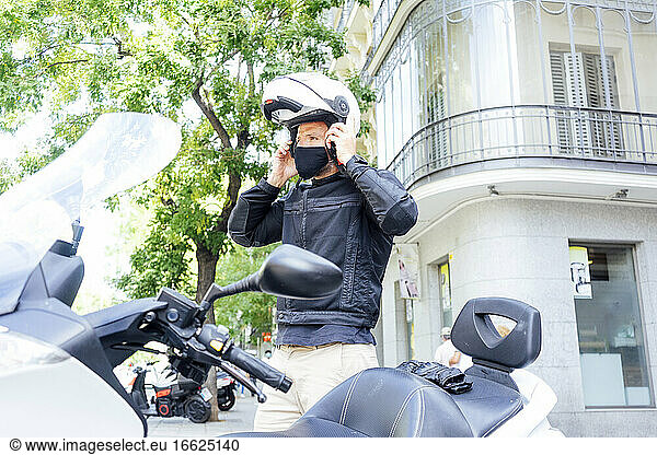 Male biker putting on helmet while standing by motor scooter