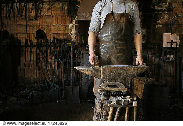 Male blacksmith standing with hammer by anvil at shop