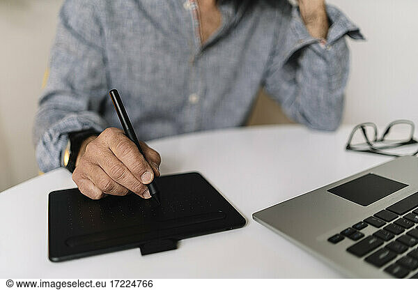 Male business professional using graphics tablet by laptop on table at home office