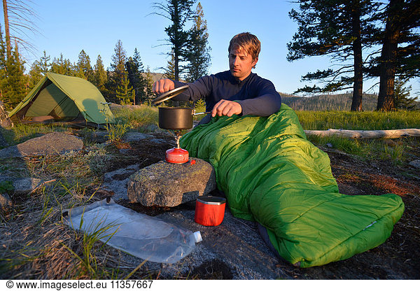 Male camper in sleeping bag prepares a meal on camping stove at Midnight Ridge  Colville National Forest  Washington State  USA