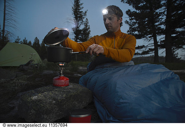 Male camper in sleeping bag prepares meal on camping stove at night on Midnight Ridge  Colville National Forest  Washington State  USA
