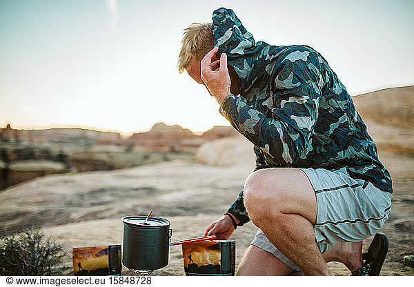 male camper prepares camp dinner and pulls camo jacket over head