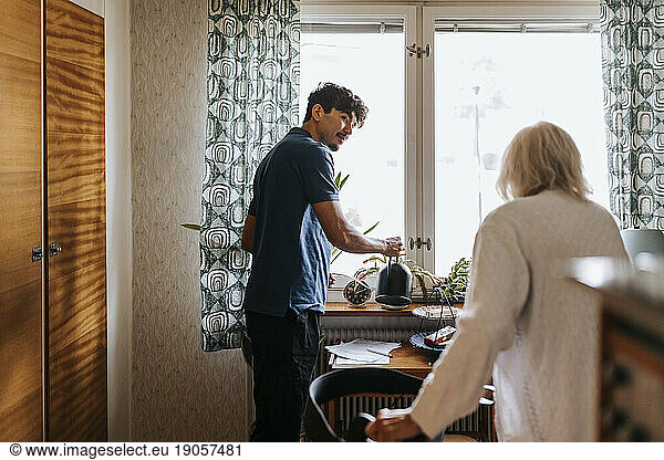 Male caregiver looking at senior woman while watering plants at home