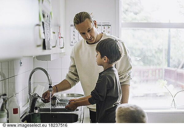 Male child care worker teaching boy to wash hands near sink at kindergarten