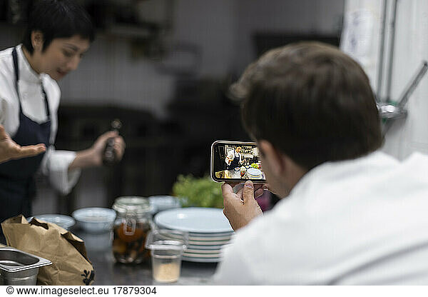 Male colleague filming chef preparing food in kitchen of restaurant